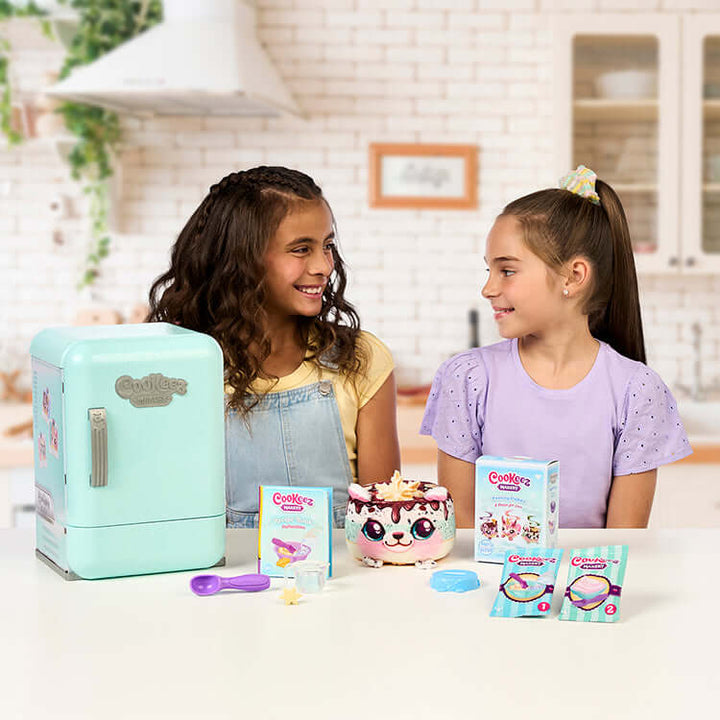 Two kids enjoying Cookeez Makery S2 Freezy Cakez Playset, featuring a mini fridge, plush cake, and playset accessories in a kitchen setting.