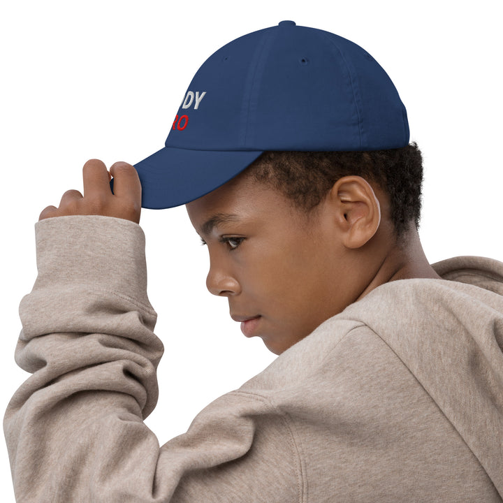 A young boy wearing an Oddy Pro Youth baseball cap, adjusting it while wearing a gray hoodie.